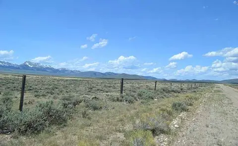 A dirt road runs alongside a fence through a landscape with distant mountains under a blue sky.
