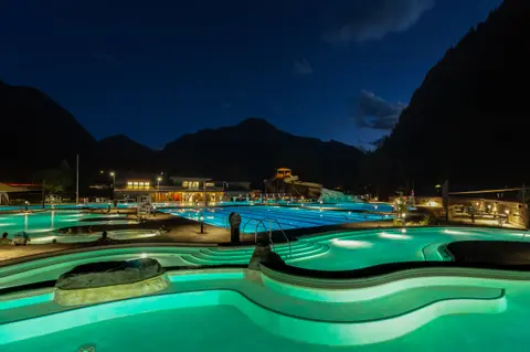 A night view of illuminated outdoor swimming pools set against dark mountains.