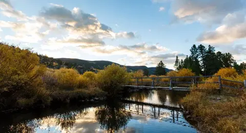 A wooden footbridge crosses a calm stream surrounded by autumn foliage and mountains under a cloudy sky.