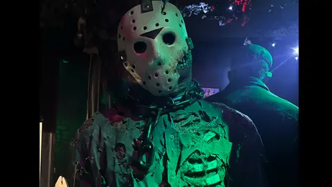 A costumed figure with a hockey mask and chains stands indoors under moody lighting.