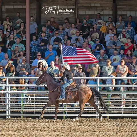A person on horseback holds an American flag in front of a seated crowd in an arena.