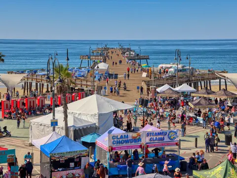 A high-angle view of a bustling pier and plaza filled with crowds, tents, and food vendors next to the ocean.
