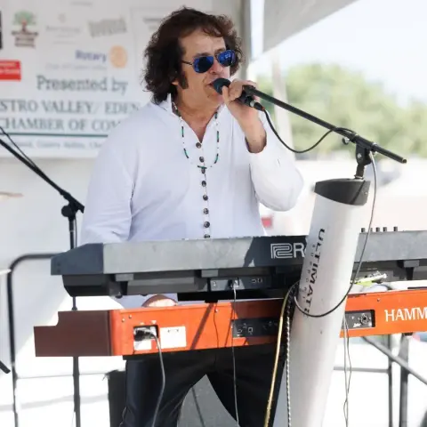 A musician performs while singing into a microphone and playing keyboards outdoors.
