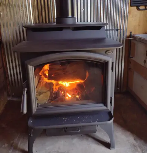 A wood-burning stove with a bright fire burning inside, set against corrugated metal wall panels.