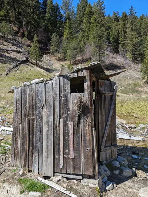 An old wooden outhouse stands in a mountainous landscape with pine trees.