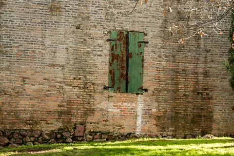 A weathered brick wall features a rusty green shuttered opening above a stone foundation and grassy ground.