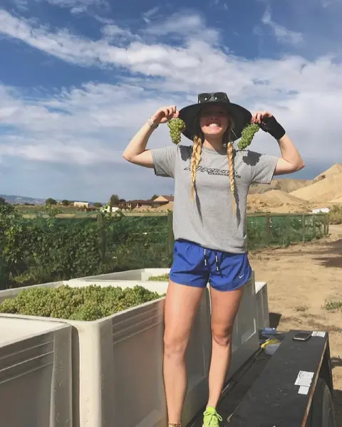 A smiling woman holding up bunches of grapes in front of bins of harvested grapes.