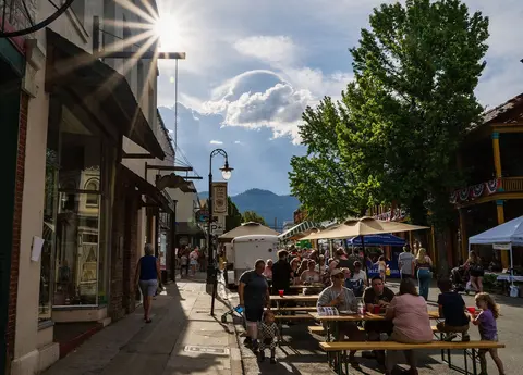 People sit at outdoor tables along a sunny street lined with historic storefronts.