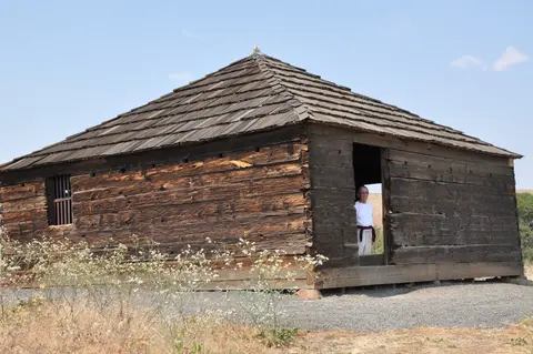 A rustic wooden building with a shingle roof and a person standing in the doorway.