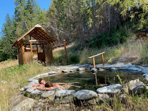 A couple relaxes in a natural stone hot spring pool outdoors near a wooden shelter and forest.