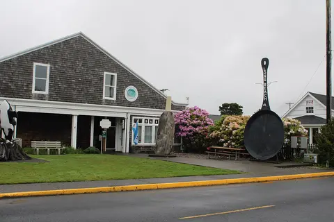 A street view of a weathered shingled building with a giant frying pan sculpture outside.