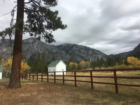 A small white building stands in a field near a wooden fence with mountains in the background.