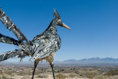 A large roadrunner sculpture made from scrap metal overlooks a valley and distant mountains.