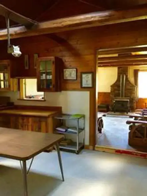 Interior view showing a rustic wooden room with a table and a doorway leading to another room with a fireplace.