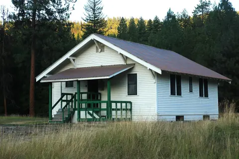 A white cabin with a brown roof and green porch stands in a forest clearing.