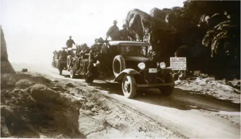 A black and white historical photo showing people riding in a truck on a dirt road.