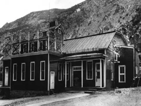 Historic black and white photograph of a dark building with a sign against a mountain backdrop.