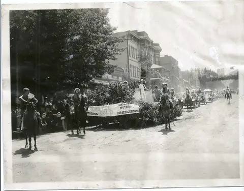 A historic photograph shows a street parade with people on horses and a decorated float.