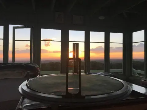 Interior view of a fire lookout tower with a fire finder and a sunset visible through the windows.