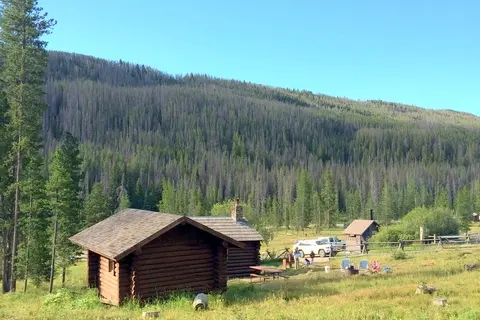 A rustic log cabin sits in a clearing in front of a heavily forested mountain slope under a clear blue sky.