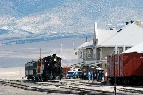 A steam train pulling into a station with buildings and mountains in the background.