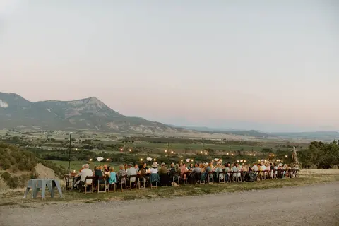 A long outdoor dinner table is set up on a hillside overlooking a valley and mountains.