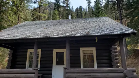 A rustic log cabin stands in a wooded area with trees and a mountain in the background.