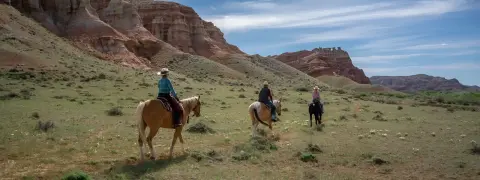 Three people ride horses across a grassy plain in front of eroded cliffs.