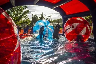 People walking inside giant inflatable balls in a pool of water under a tent.