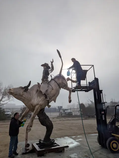Two workers clean and maintain a large statue of a cowboy riding a bucking bull.