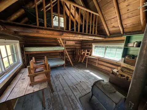 The interior of a rustic log cabin features bunk beds, a loft, a wood stove, and wooden furniture.