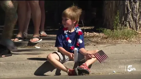 A young boy sitting on a curb holding an American flag and wearing a patriotic lei.