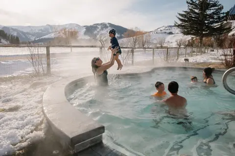 A family enjoys an outdoor hot spring pool with snow-covered mountains in the background.