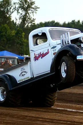 A white vintage pickup truck is pulling with its front wheels off the ground on a dirt track.