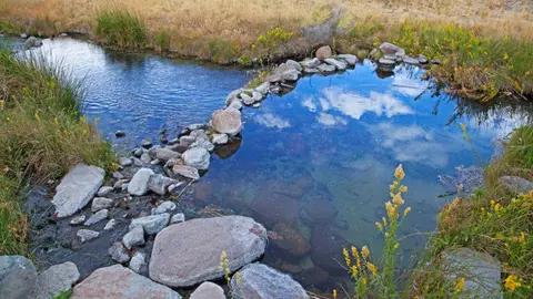 A natural hot spring pool bordered by rocks and grassy banks with reflections of the sky.