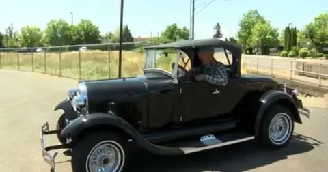 A person drives a vintage black convertible car outdoors.