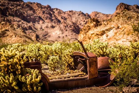 An old rusted car body is surrounded by desert cactus and rocky hills.