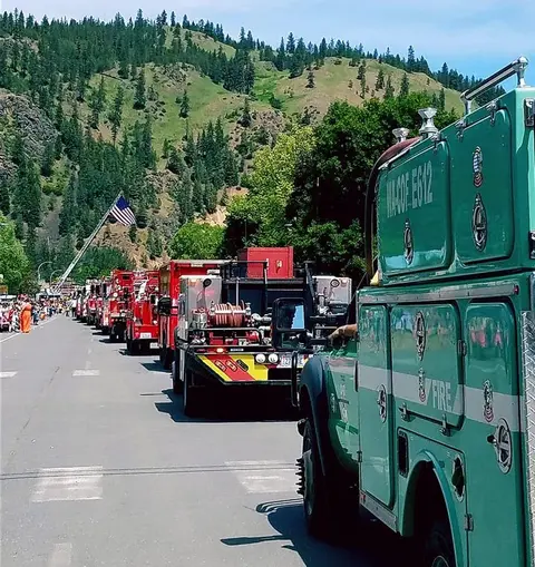 A line of fire trucks parked on a street with a hill and flag in the background.