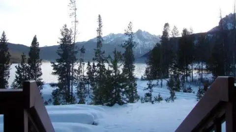 A snowy landscape with trees, water, and mountains in the distance viewed from a porch.