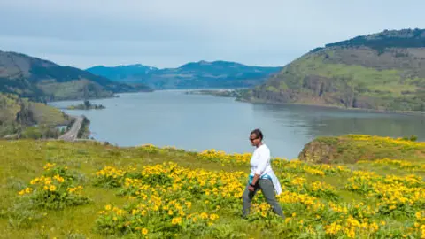 A person walks through a field of yellow wildflowers overlooking a large river and hills.