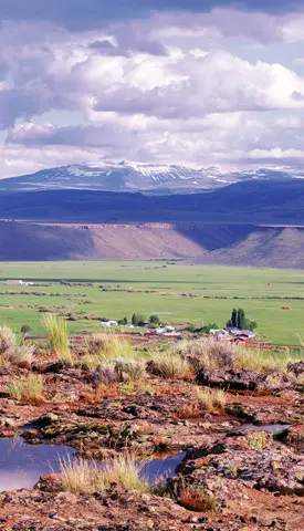 A scenic landscape showing a wide green valley with distant mountains and a cloudy sky.