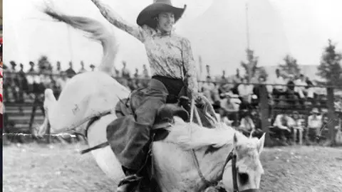 Historic black and white photograph of a cowgirl riding a bucking horse at a rodeo.
