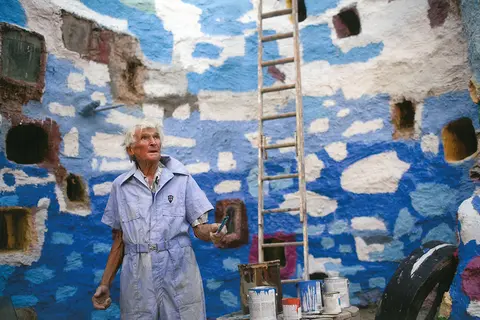 An elderly man in a blue jumpsuit stands inside a painted stone structure with a ladder and paint cans.