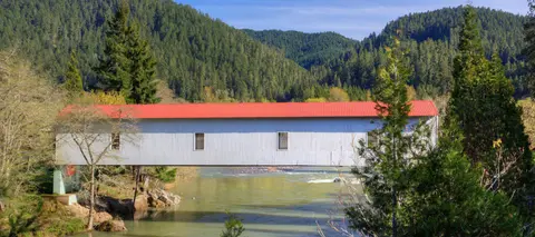 A covered bridge spans a river against a backdrop of forested hills.