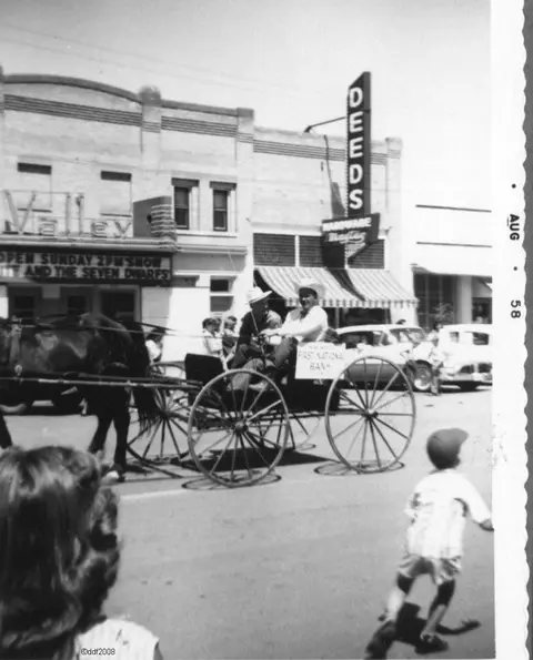 Black and white historic photo showing people in a horse-drawn carriage on a street.