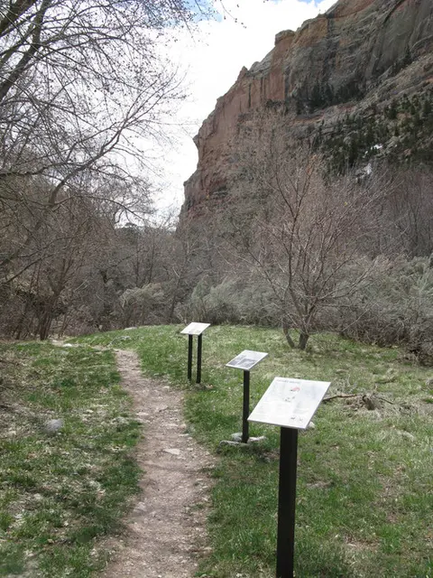 A dirt path leads past several informational signs in a landscape with cliffs and sparse trees.