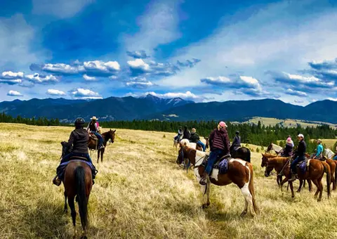 A group of people ride horses across a grassy field with mountains in the distance.