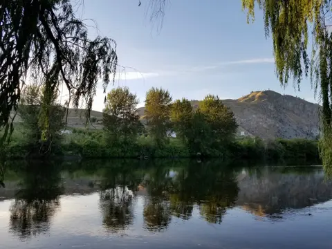 A calm river reflecting trees and a hill under a clear sky.