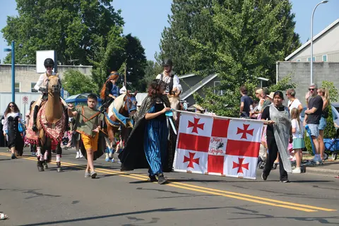 People dressed in medieval costumes walk in a street parade carrying a large banner and leading horses.
