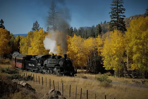 A steam train travels through a landscape with autumn-colored trees.
